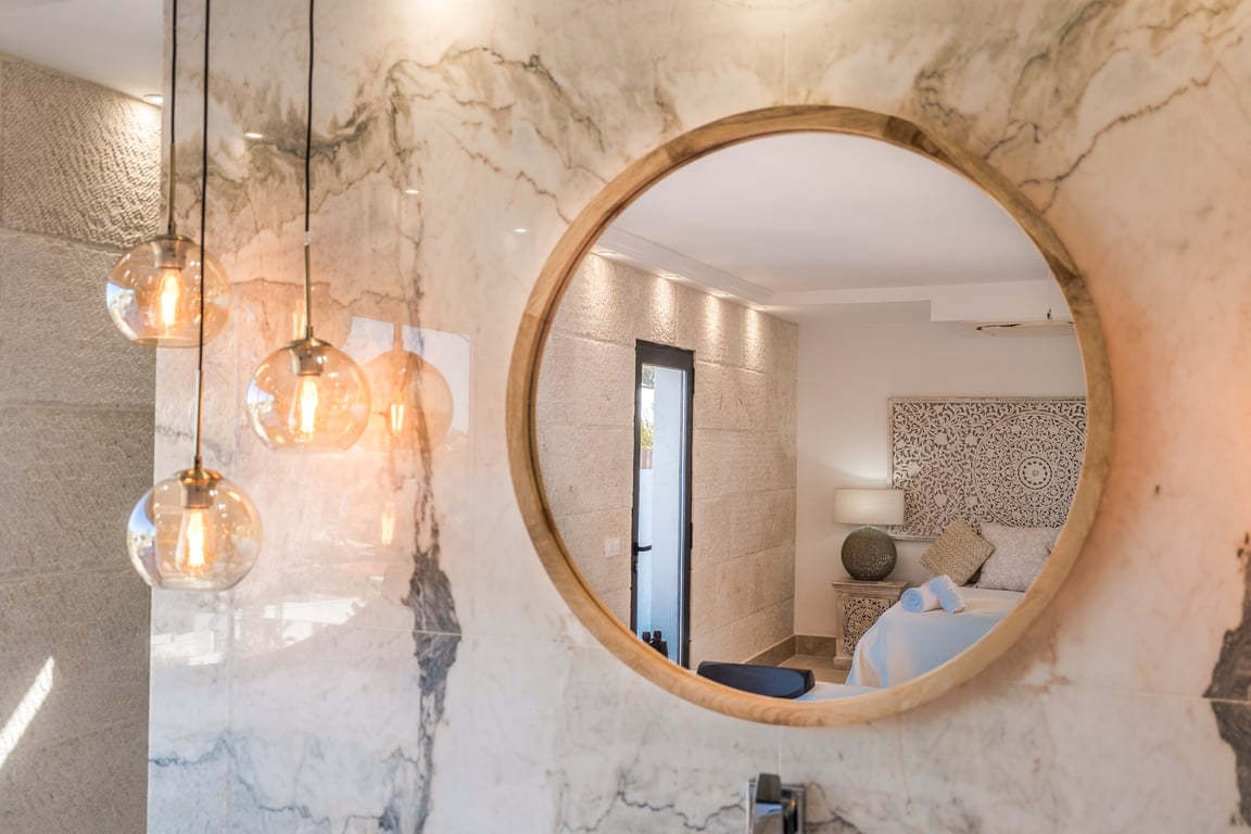 Espejo circular con marco de madera sobre pared de mármol veteado, iluminado por lámparas colgantes de cristal y reflejo del dormitorio con muros de piedra.