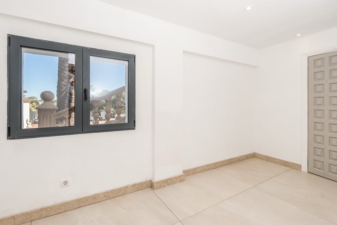 Habitación renovada con suelos de piedra natural, focos empotrados y ventana de aluminio oscuro con vistas a la zona exterior de la villa.