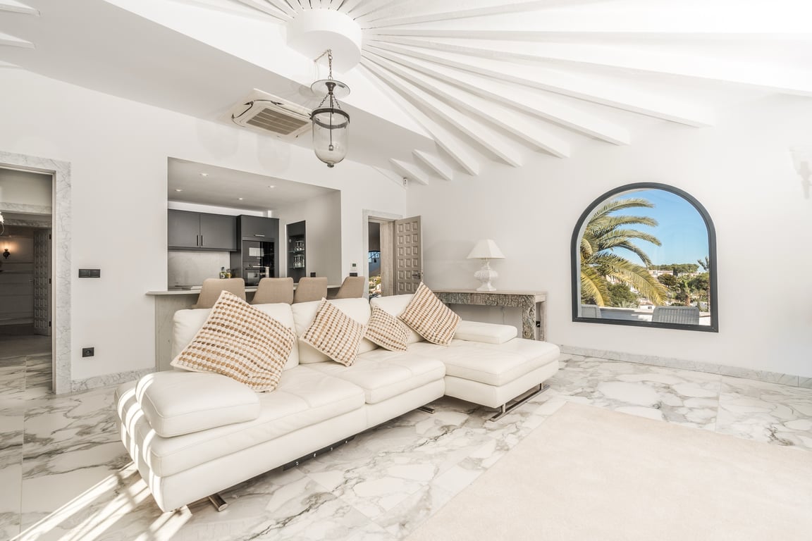 Salón luminoso con suelos de mármol, sofá blanco y un techo radial único. La estancia conecta con la cocina semiabierta y ofrece vistas al exterior por un ventanal en arco.