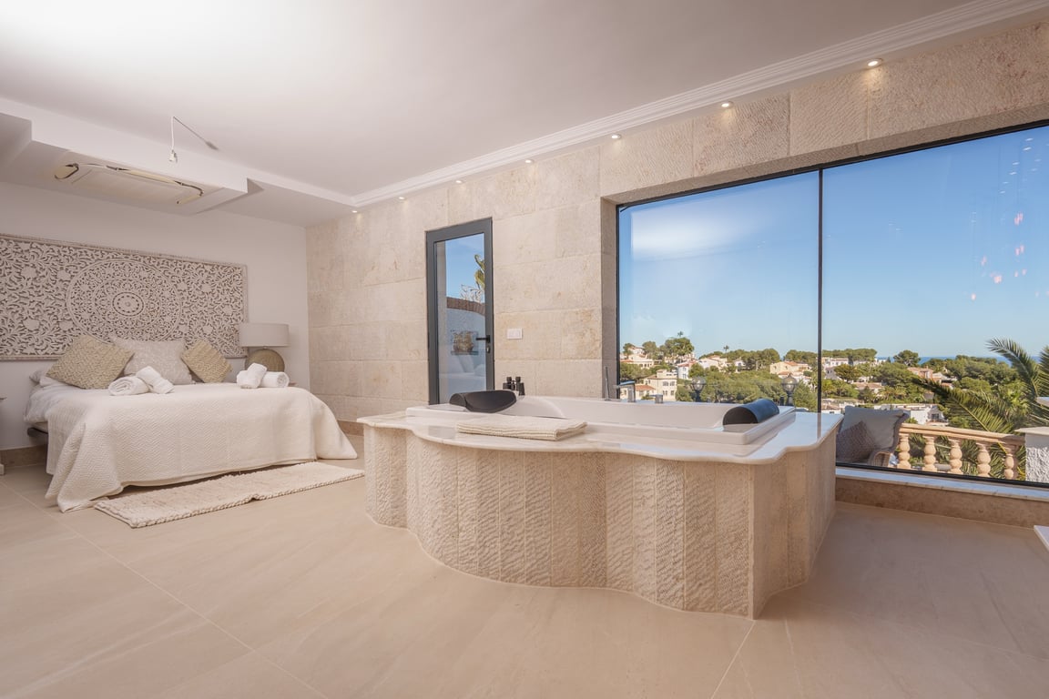 Suite principal con bañera integrada revestida en piedra natural, amplios ventanales con vistas despejadas al mar y una decoración en tonos serenos.