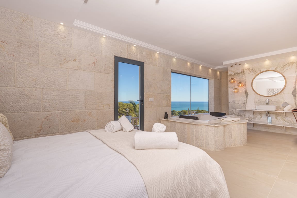 Suite principal con paredes de piedra natural, jacuzzi integrado junto al ventanal con vistas al mar y baño de concepto abierto con acabados en mármol.