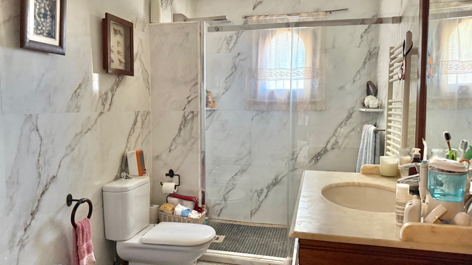Baño con paredes revestidas en mármol, ducha de obra con mampara de cristal y encimera de mármol. Incluye radiador toallero y una pequeña ventana de arco.
