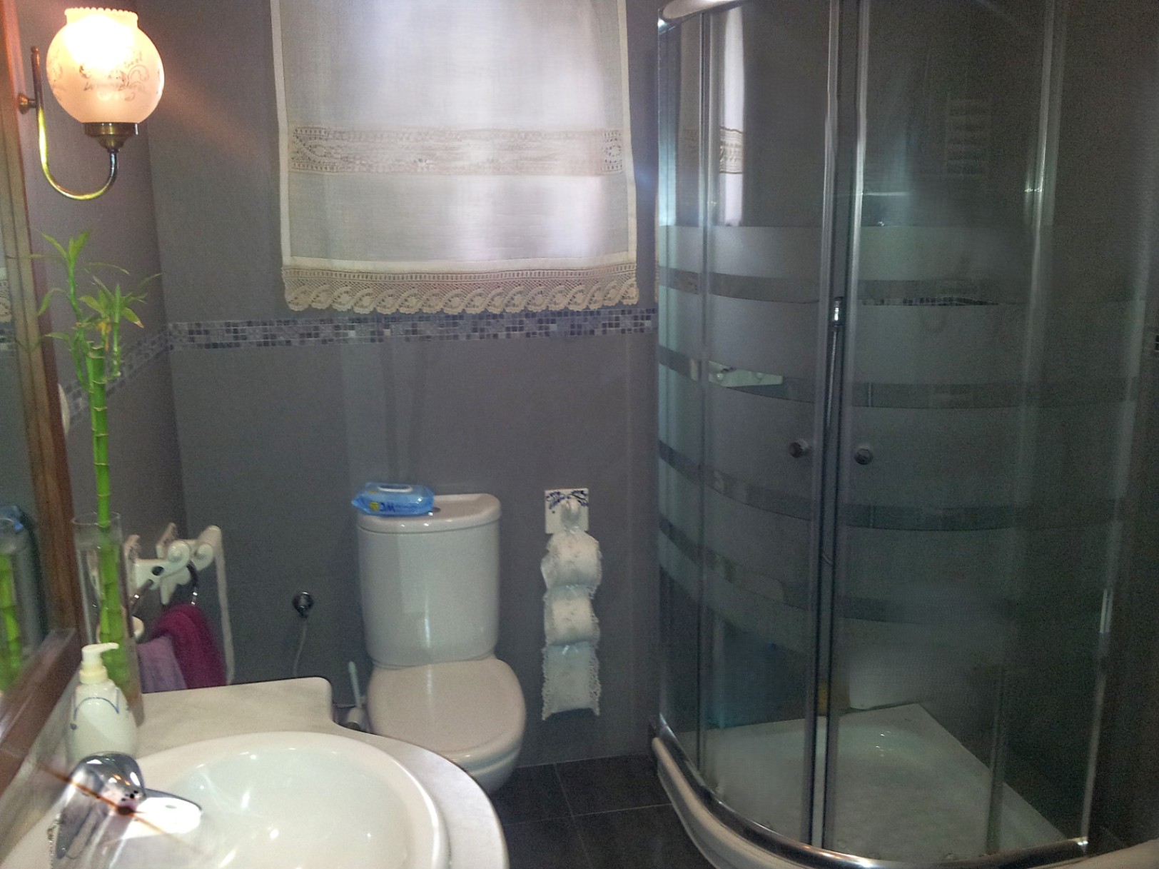 Baño funcional con cabina de ducha de esquina, encimera de mármol y paredes revestidas en tonos grises con cenefa de mosaico.