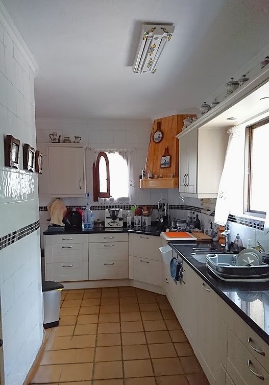 Cocina independiente de estilo tradicional con armarios blancos, encimeras oscuras y suelos de terracota. Incluye una campana revestida de madera y paredes alicatadas con cenefa decorativa.
