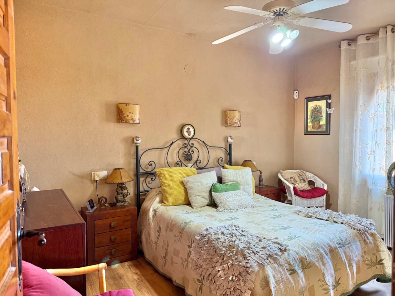 Dormitorio con cabecero de forja, mesitas de noche de madera y ventilador de techo. Incluye un sillón de mimbre y luz natural a través de ventanales con cortinas.