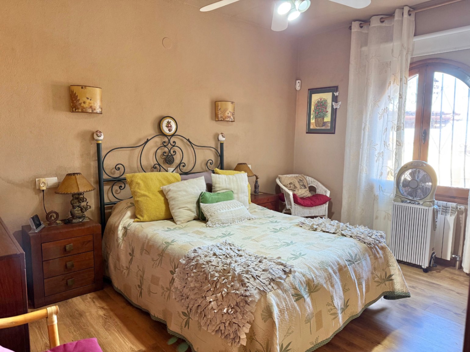 Dormitorio con cabecero de forja, suelos de madera y ventana de arco. Incluye ventilador de techo, mobiliario rústico y calefacción por radiador.