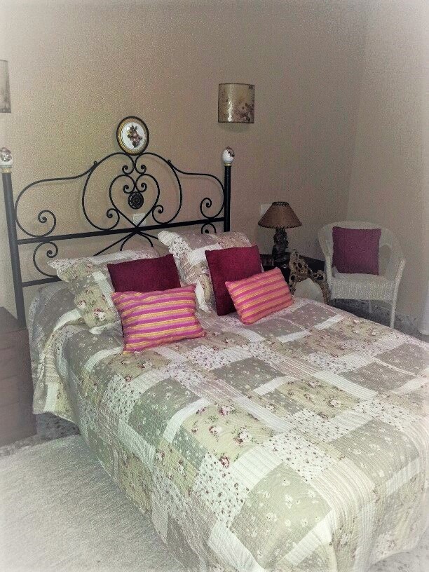 Dormitorio con cabecero de hierro forjado negro, colcha de patchwork y cojines decorativos. Incluye un sillón de mimbre blanco y apliques de pared de estilo clásico.