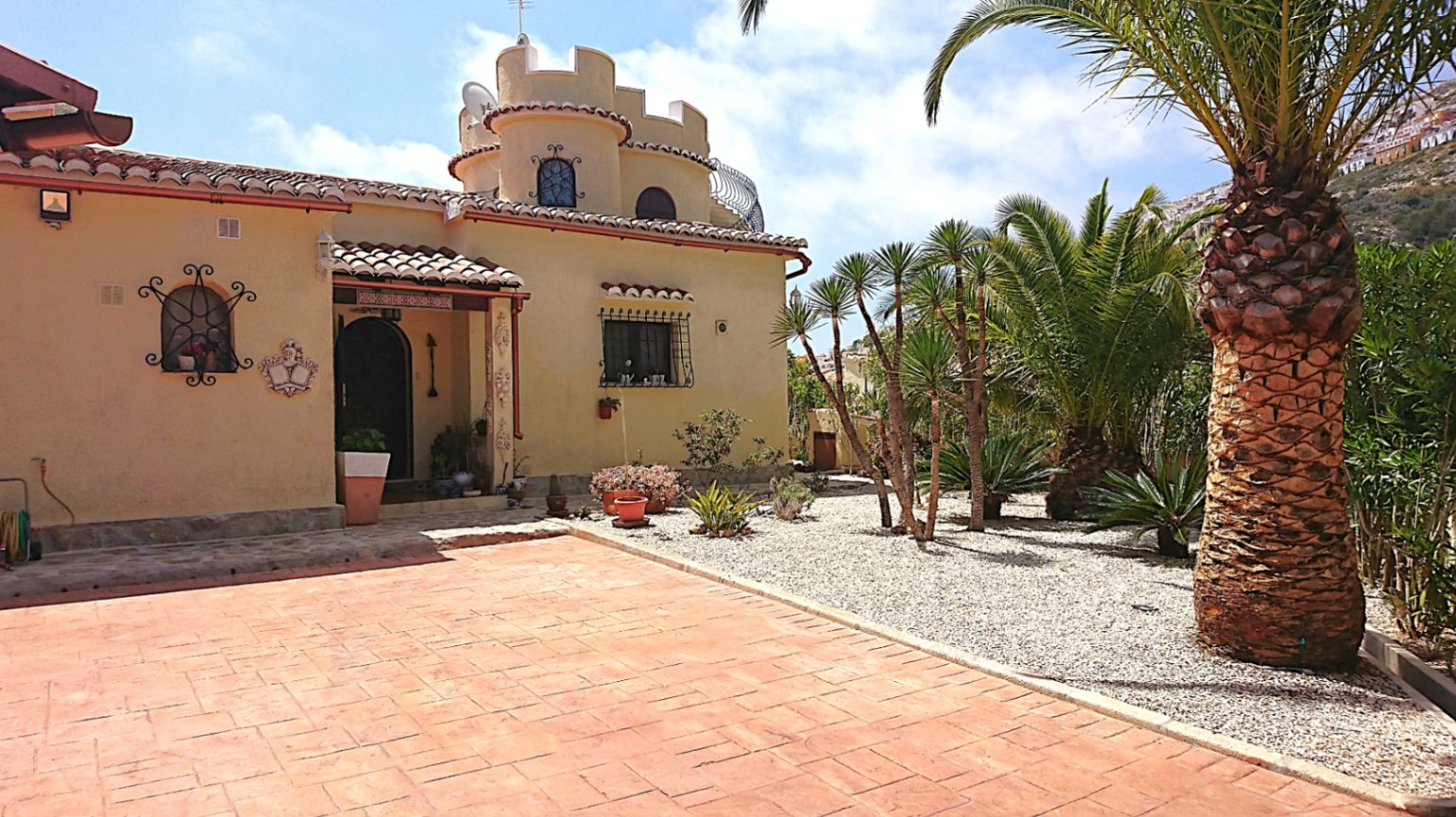 Fachada principal con torre circular almenada, ventanas de arco con rejería y jardín mediterráneo con palmeras y suelo de hormigón impreso.