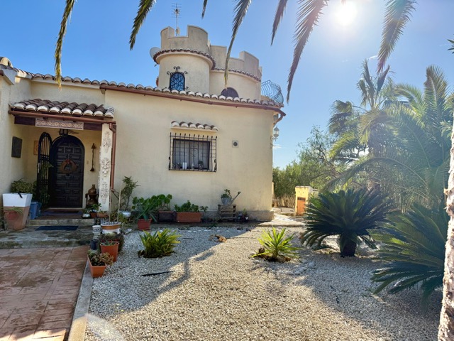 Fachada principal de la villa con su característica torre circular, porche de entrada con teja árabe y jardín mediterráneo de grava con palmeras.