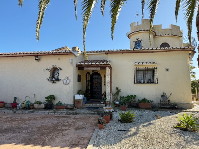Fachada principal de la villa con torre circular almenada, carpintería de madera oscura y rejas de hierro forjado sobre paredes de estuco crema.