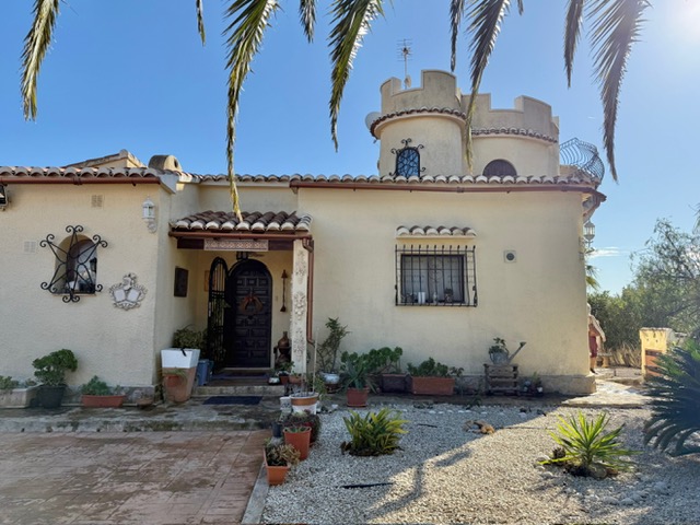 Fachada principal de la villa con torre circular almenada, entrada de madera tallada y detalles de forja en ventanas, rodeada de un jardín de grava y plantas mediterráneas.