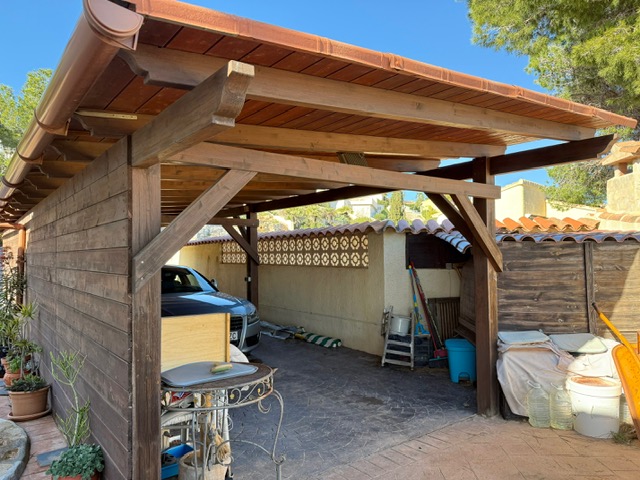 Porche cubierto de estructura de madera con tejado de teja cerámica y suelo de hormigón impreso, diseñado para el estacionamiento de vehículos en la parcela.