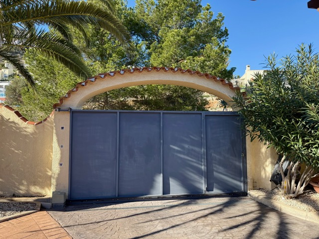 Puerta de entrada corredera bajo un arco de estilo mediterráneo con remate de teja, rodeada de vegetación madura y acceso pavimentado.