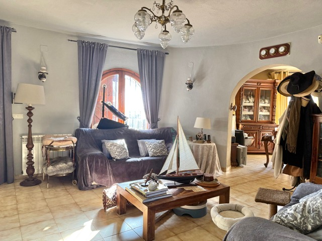 Salón de planta circular con suelos cerámicos, ventana de madera en arco y acceso mediante arco al comedor, decorado con elementos náuticos y luz natural.