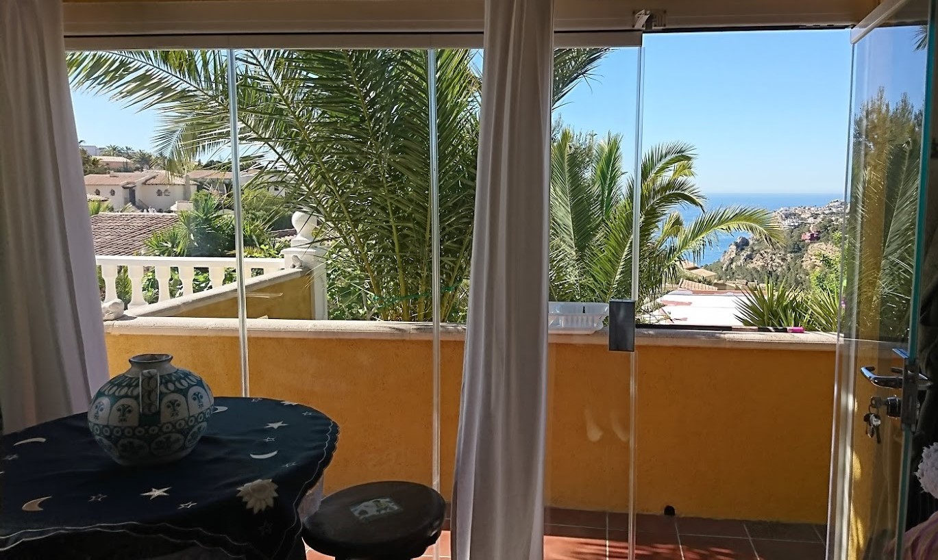 Terraza acristalada con cortinas de cristal sin marcos, ofreciendo vistas al mar Mediterráneo y al jardín con palmeras maduras.