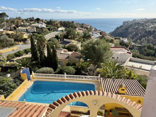 Vista elevada de la piscina rectangular con balaustrada blanca, enmarcada por arcos de teja y vistas panorámicas al mar Mediterráneo y la costa de Benitachell.