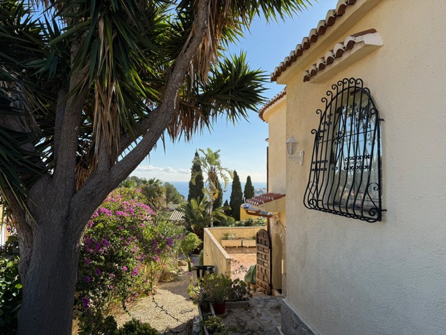Vista exterior de la villa con fachada mediterránea, rejas de hierro forjado y jardín maduro con buganvillas que enmarca las vistas al mar.
