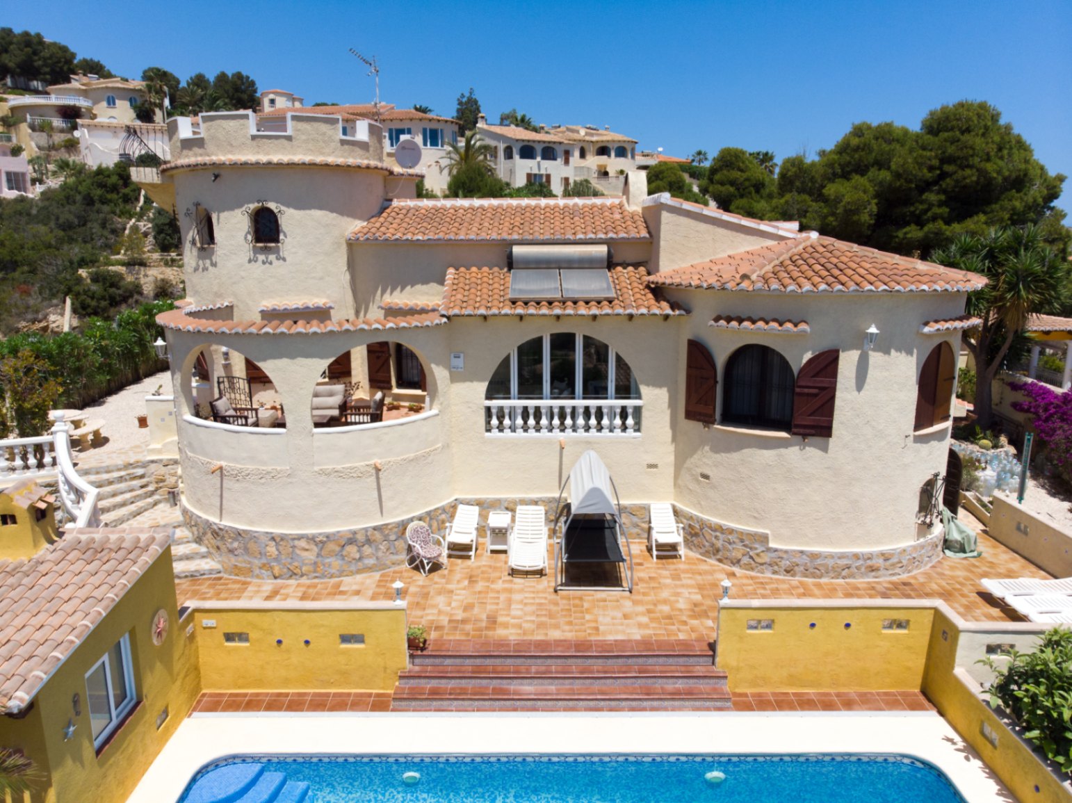 Vista exterior de la villa con torre circular, piscina privada, amplias terrazas soleadas y paneles solares sobre el tejado de teja tradicional.