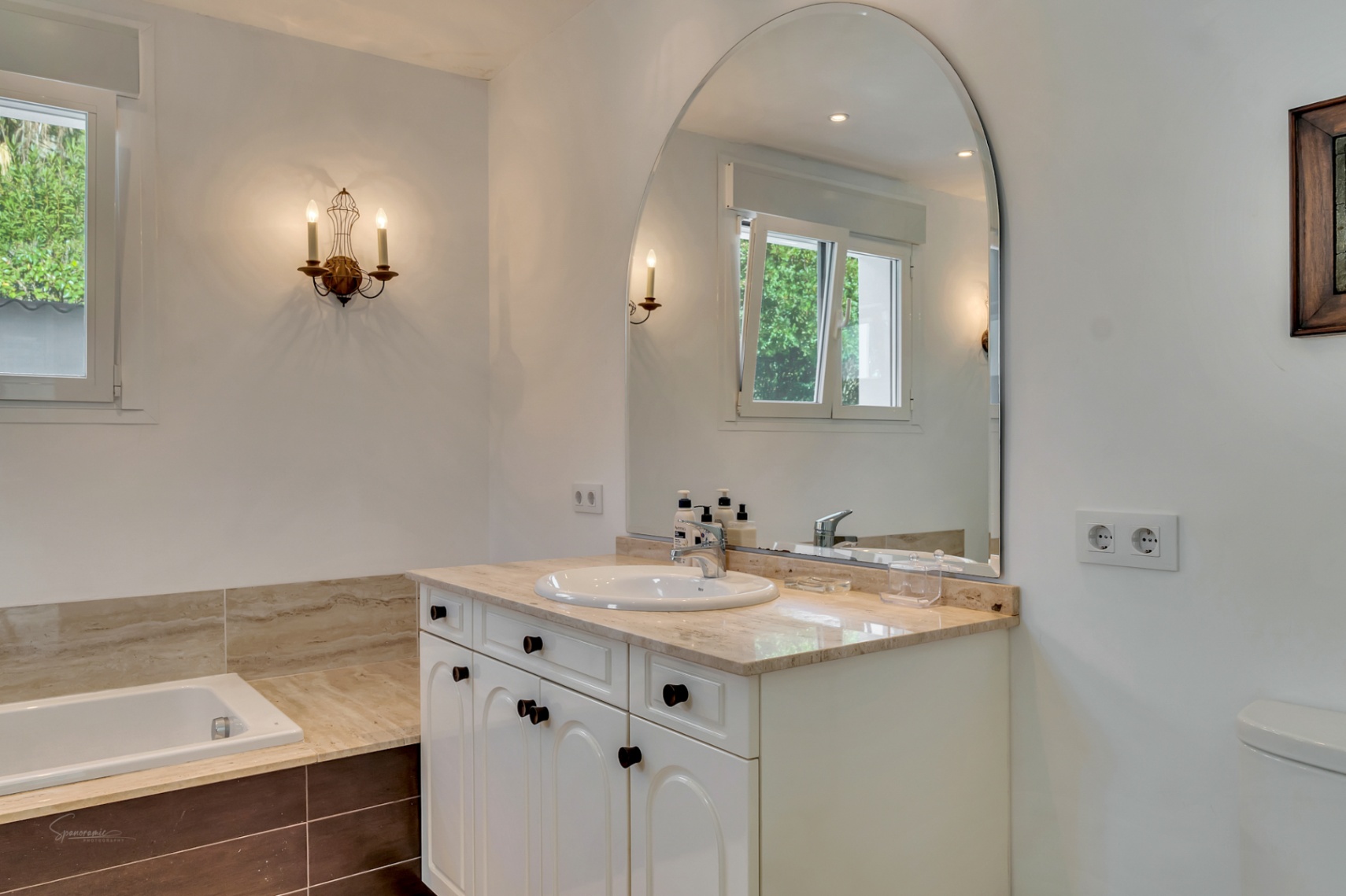 Baño elegante con encimera de mármol, lavabo integrado y un gran espejo de arco. Incluye bañera encastrada con acabados en piedra y apliques de pared de estilo clásico.