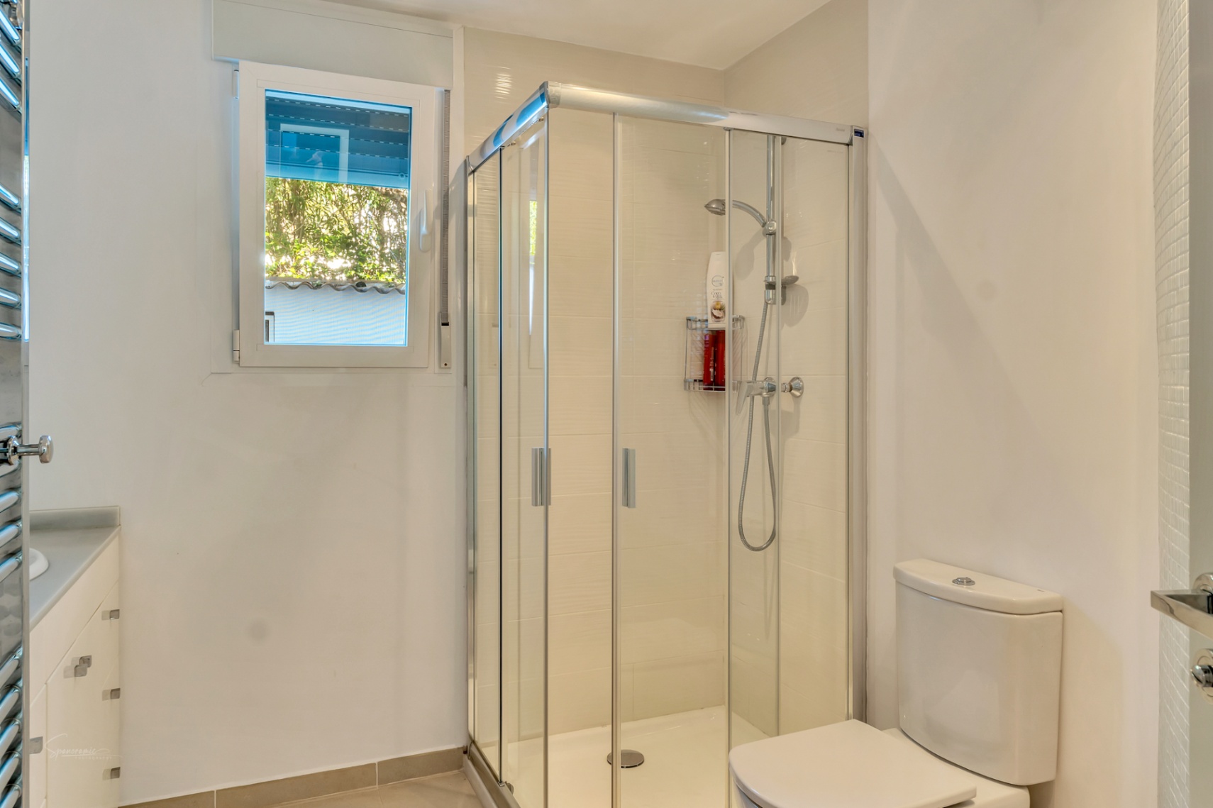 Baño moderno con cabina de ducha acristalada, sanitarios blancos y ventana exterior que aporta luz natural. Incluye un mueble lavabo con cajoneras y toallero radiador cromado.