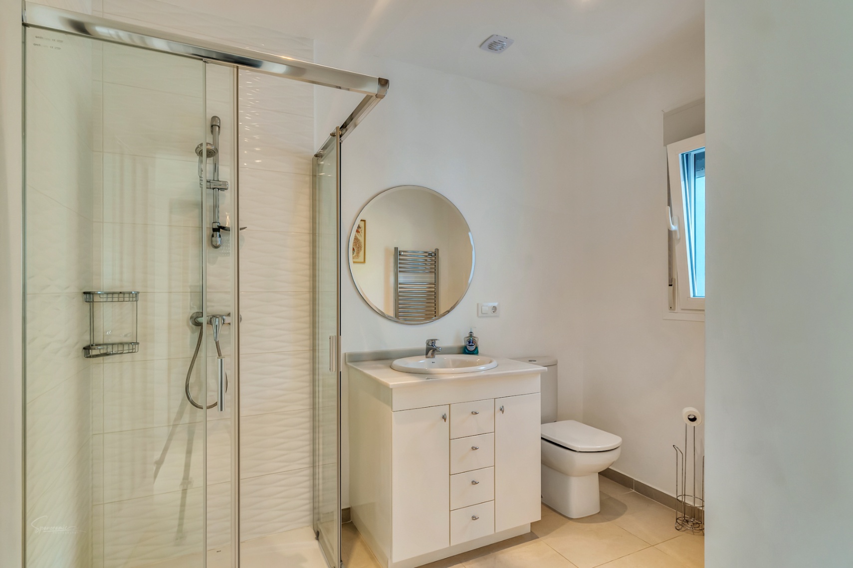 Baño moderno con ducha de obra acristalada, azulejos con relieve, mueble de lavabo blanco con espejo circular y luz natural desde una ventana lateral.