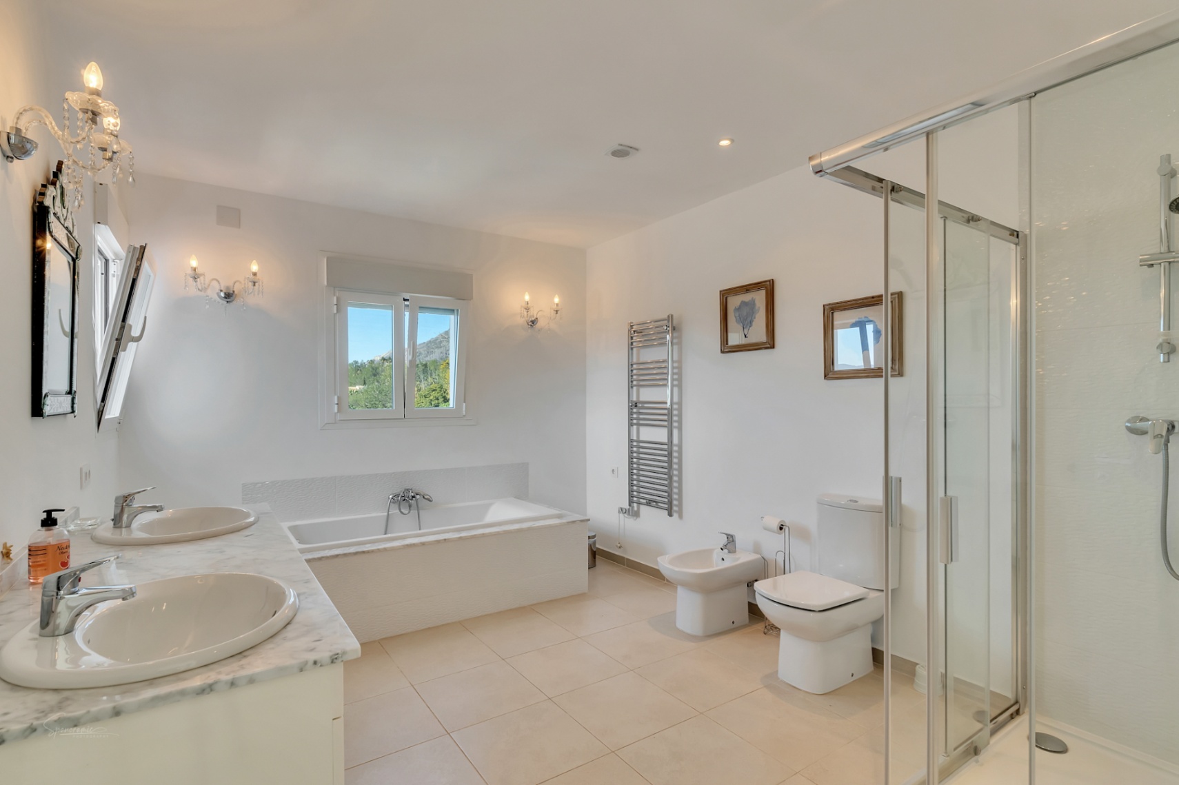 Baño principal con encimera de mármol y lavabo doble, bañera integrada bajo ventana con vistas al Montgó, ducha acristalada y elegantes apliques de pared tipo candelabro.