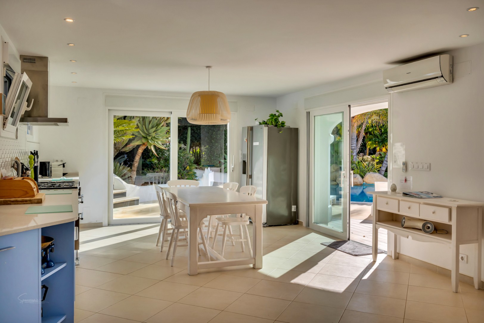 Cocina abierta y comedor con mesa blanca, muebles azules y acceso directo a la piscina y jardín mediante puertas correderas, con iluminación empotrada y aire acondicionado.
