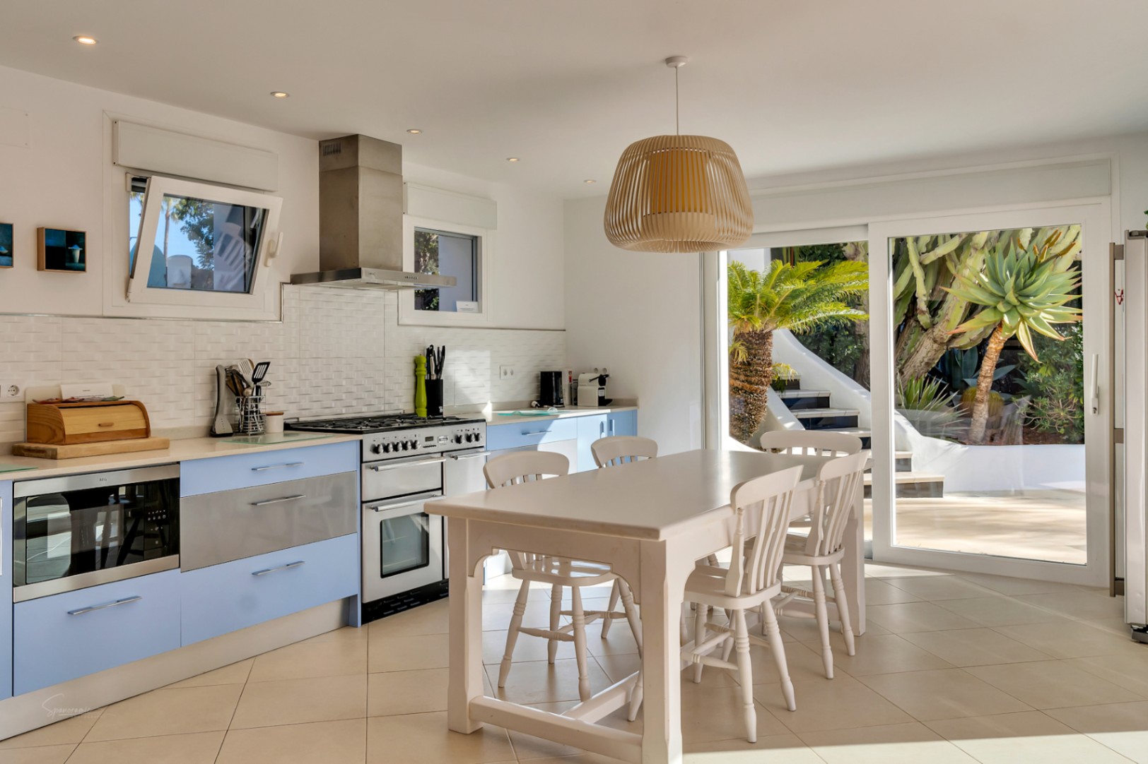 Cocina de diseño mediterráneo con armarios azul pastel, encimera blanca y zona de comedor integrada. Las amplias puertas correderas de cristal conectan el interior con el jardín.