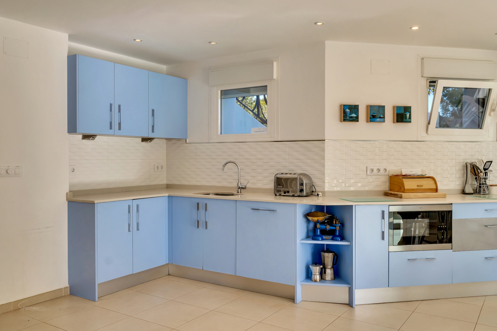 Cocina moderna con armarios en azul pastel, encimera clara y salpicadero de azulejos blancos. Incluye electrodomésticos integrados y ventanas que aportan luz natural.