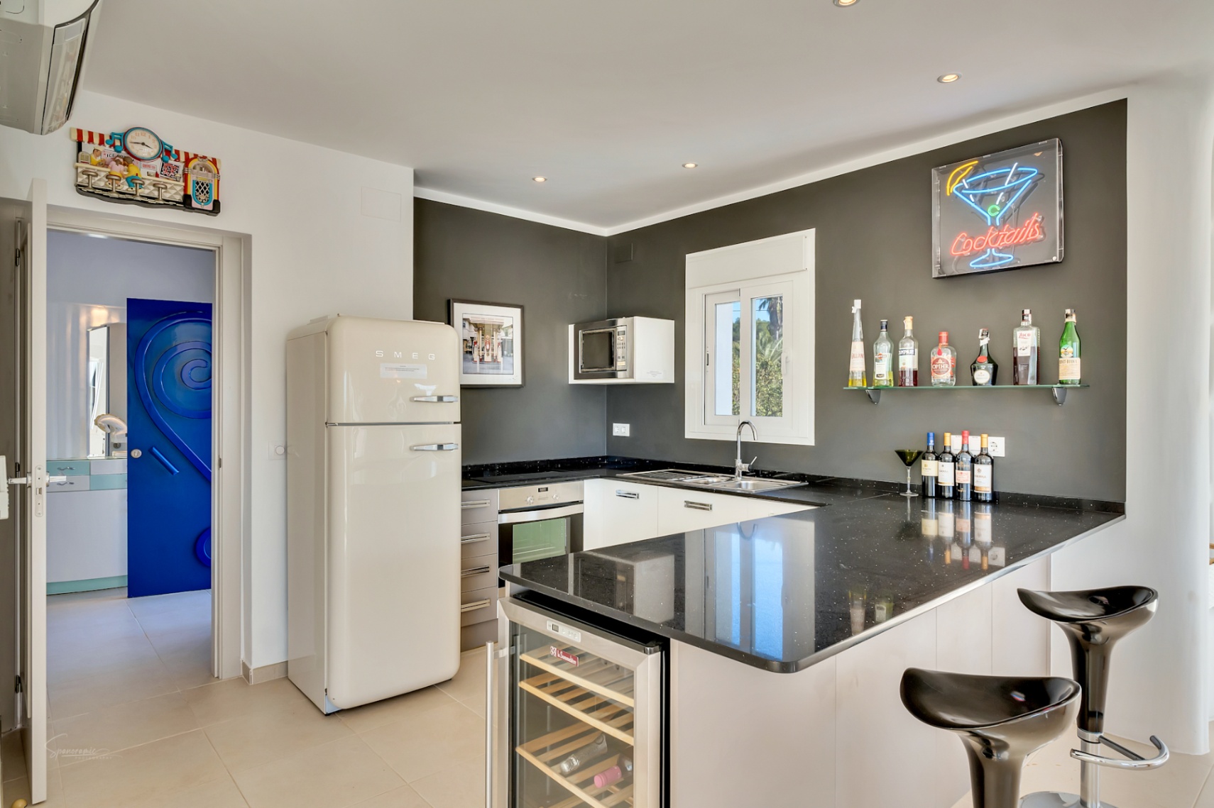 Cocina moderna con encimera de granito negro, nevera Smeg estilo retro y barra con vinoteca integrada. Destaca la pared gris con estantes de cristal y un letrero de neón.