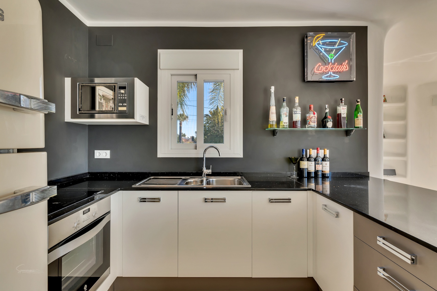 Cocina moderna con encimera de granito negro, paredes en gris antracita y una zona de bar integrada con estante de cristal y cartel de neón.
