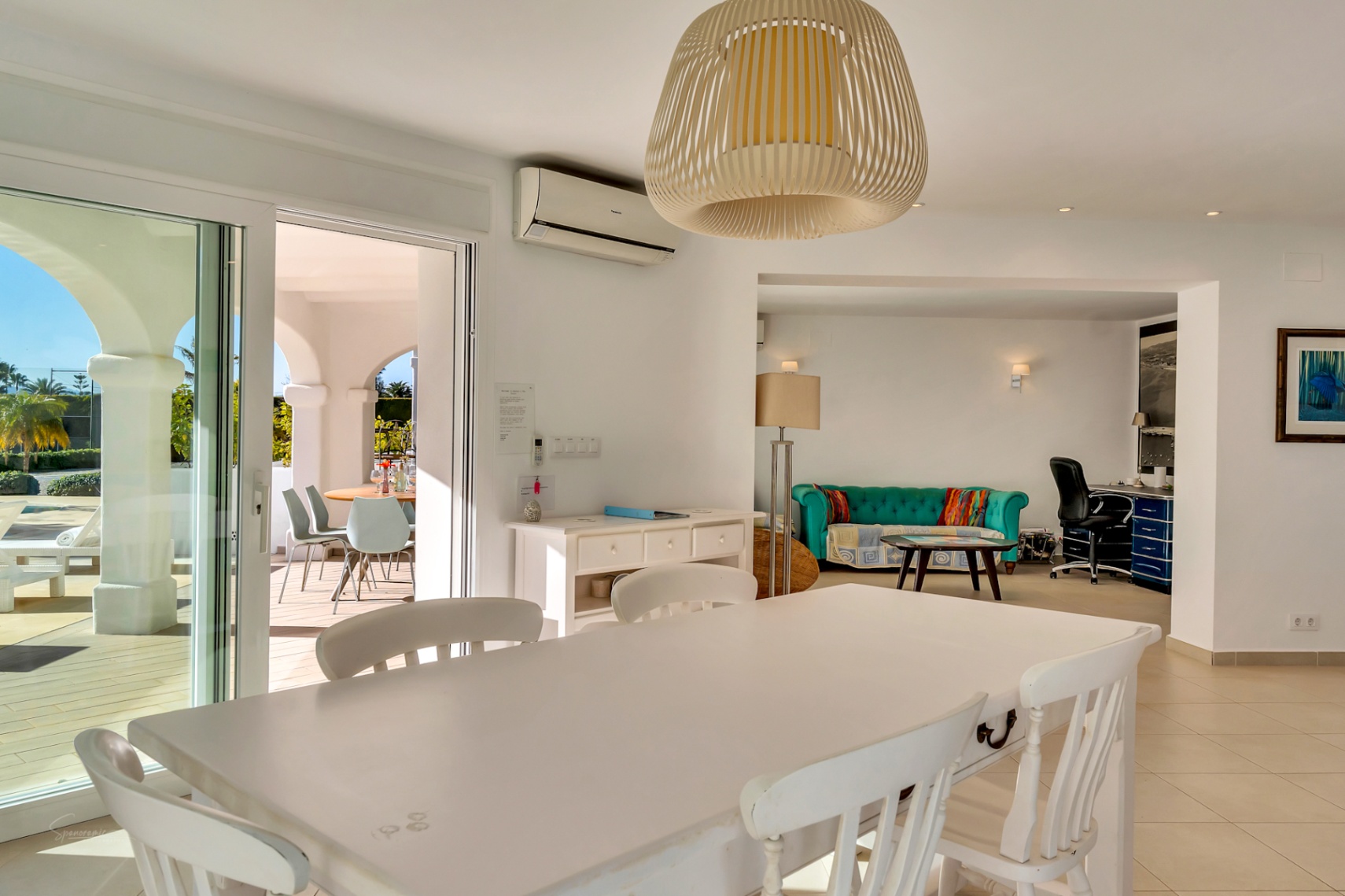 Comedor luminoso con mesa de madera blanca y lámpara de mimbre, conectado a una naya con arcos y un área de estar con despacho integrado.