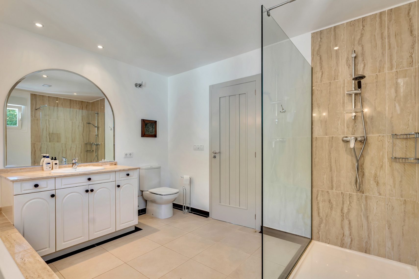 Cuarto de baño elegante con ducha a ras de suelo revestida en piedra natural, lavabo con encimera de mármol y un gran espejo de arco que amplía el espacio.