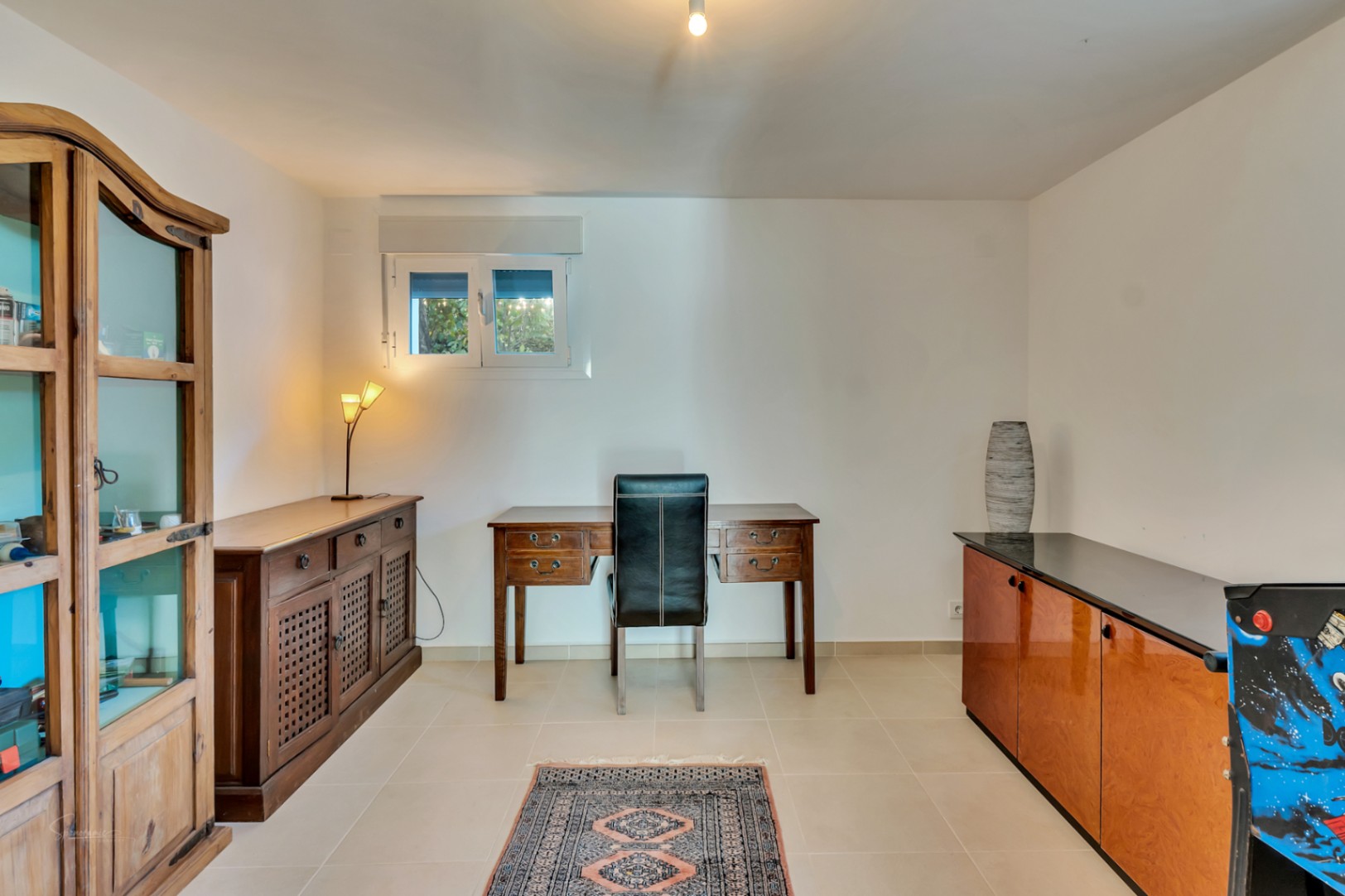 Despacho funcional con escritorio de madera clásica, silla de cuero negro y mobiliario rústico. Incluye una vitrina de madera y suelos cerámicos claros bajo luz natural.