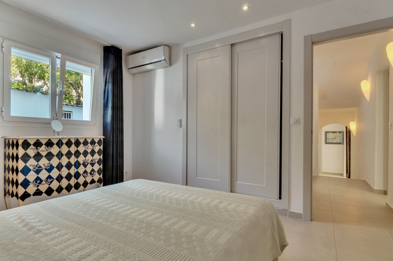Dormitorio con armarios empotrados, aire acondicionado y una cómoda de diseño arlequín. La ventana ofrece luz natural y vistas directas al jardín exterior.