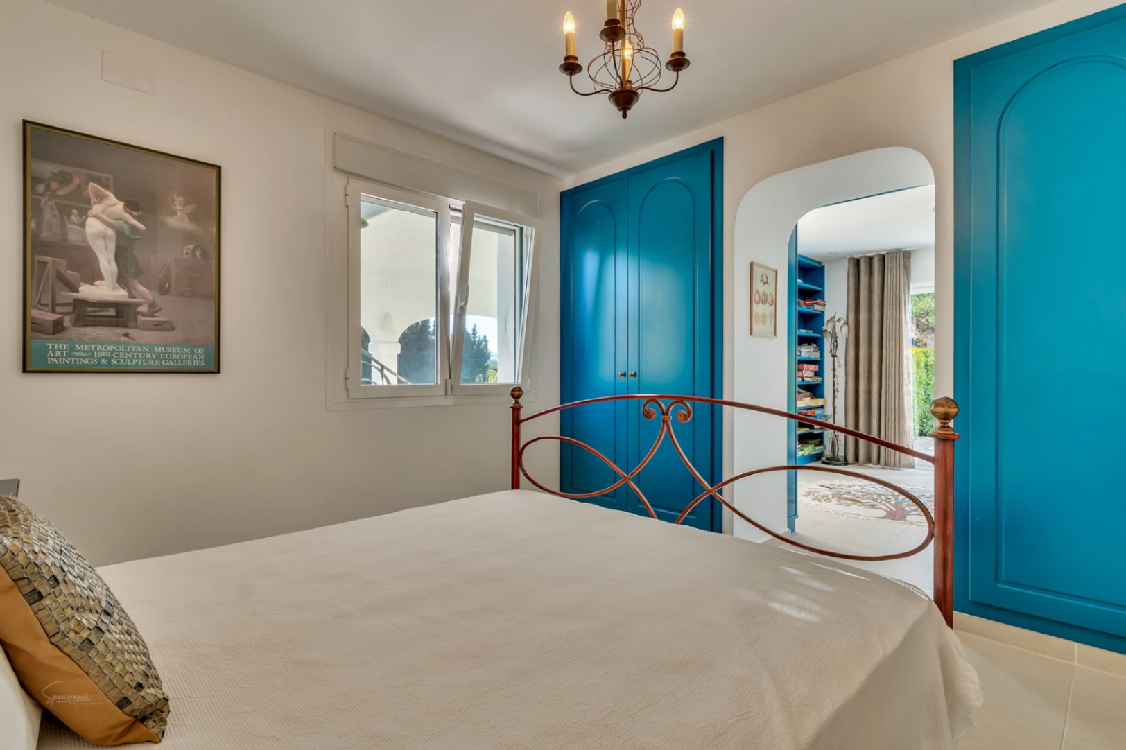 Dormitorio con armarios empotrados en azul vibrante, cabecero de hierro forjado y acceso a un estudio privado a través de un elegante arco.