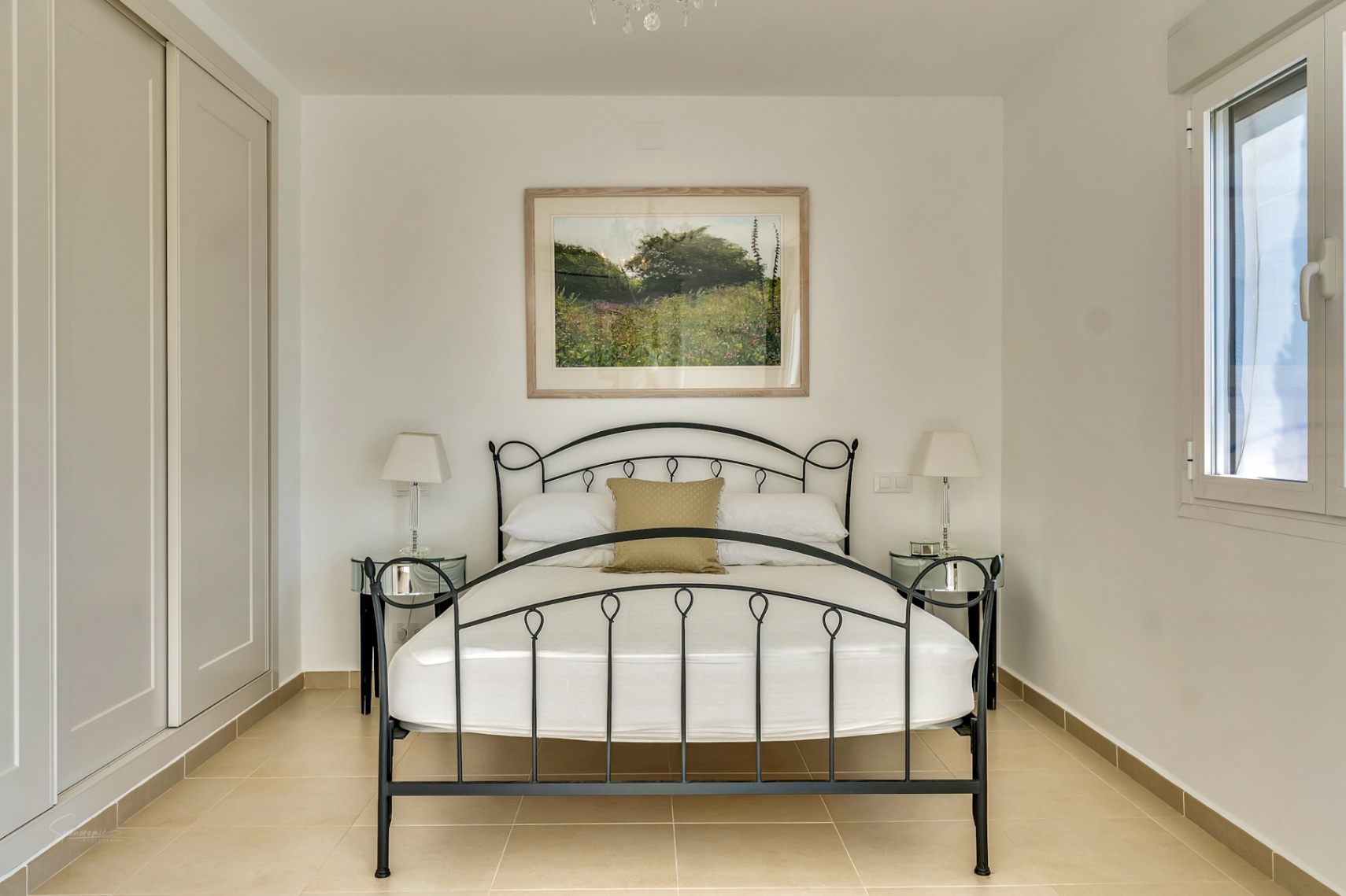 Dormitorio con cama de forja negra, armarios empotrados de color crema, suelos de baldosa clara y una ventana lateral que aporta luz natural.