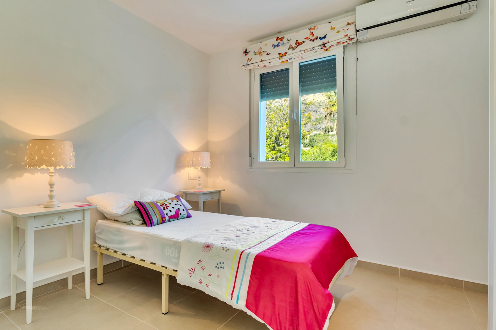 Dormitorio individual luminoso con aire acondicionado split, suelos cerámicos y ventana con vistas directas al jardín y al macizo del Montgó.