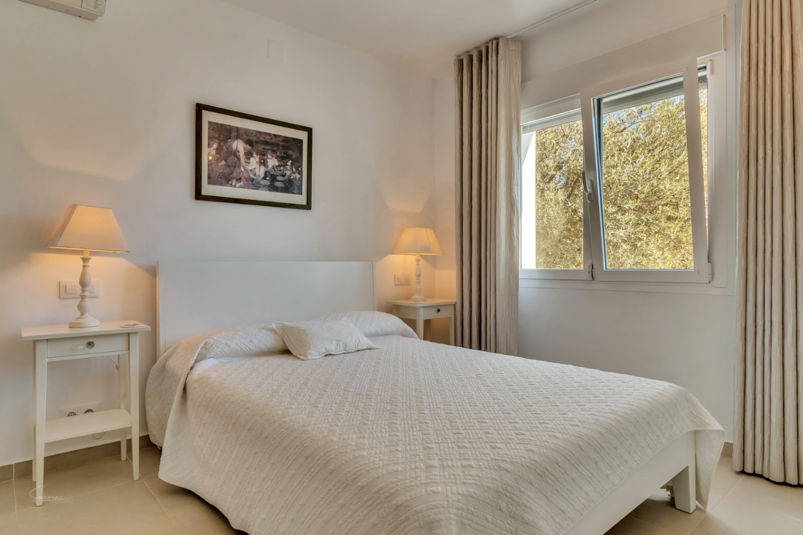 Dormitorio luminoso con cama blanca, mesitas de noche coordinadas y lámparas de luz cálida. La ventana ofrece vistas a la vegetación exterior bajo cortinas de tonos neutros.