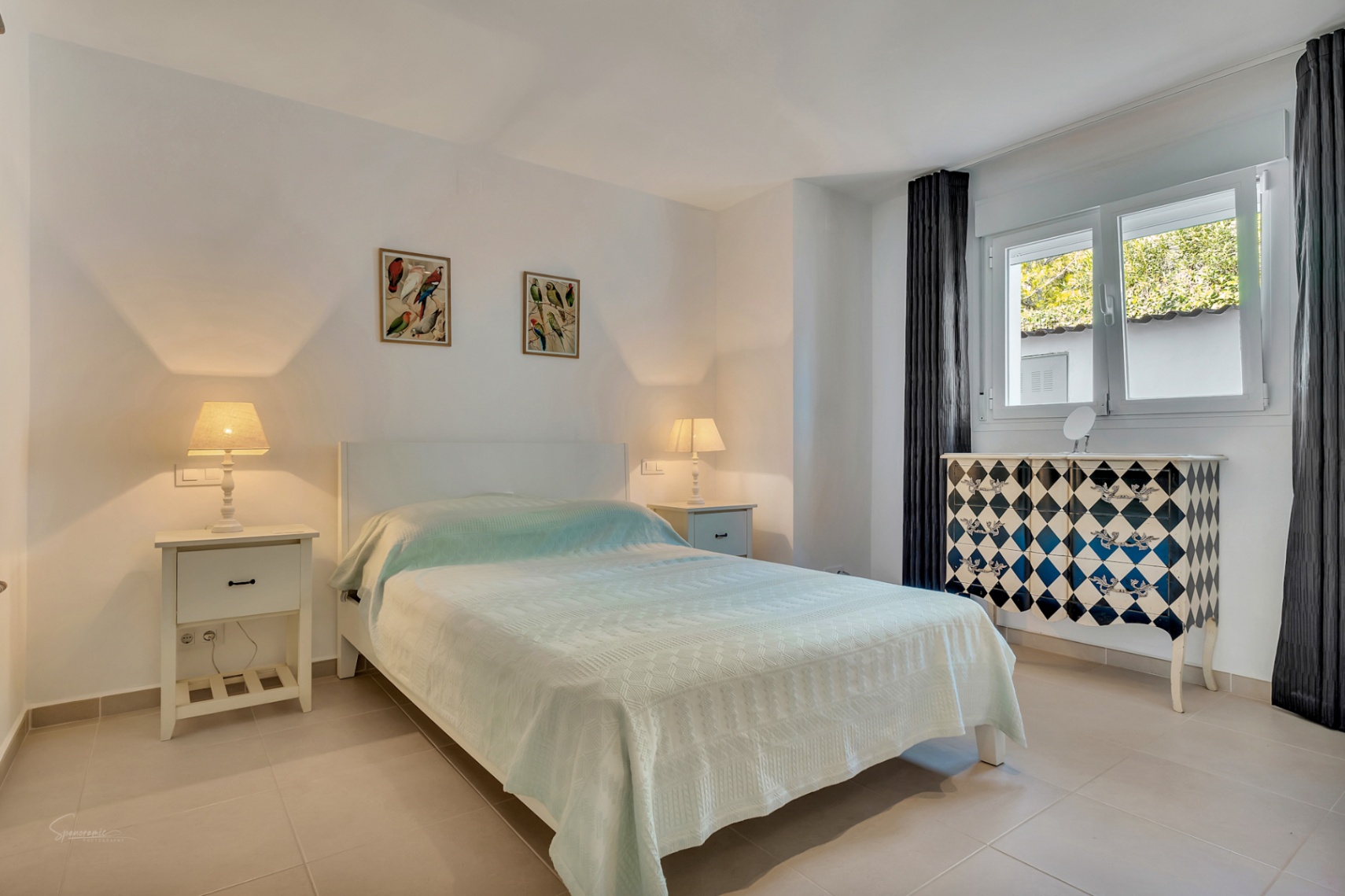 Dormitorio luminoso con suelos cerámicos, cama doble, mesitas de noche blancas y una cómoda decorativa con patrón de rombos junto a la ventana.