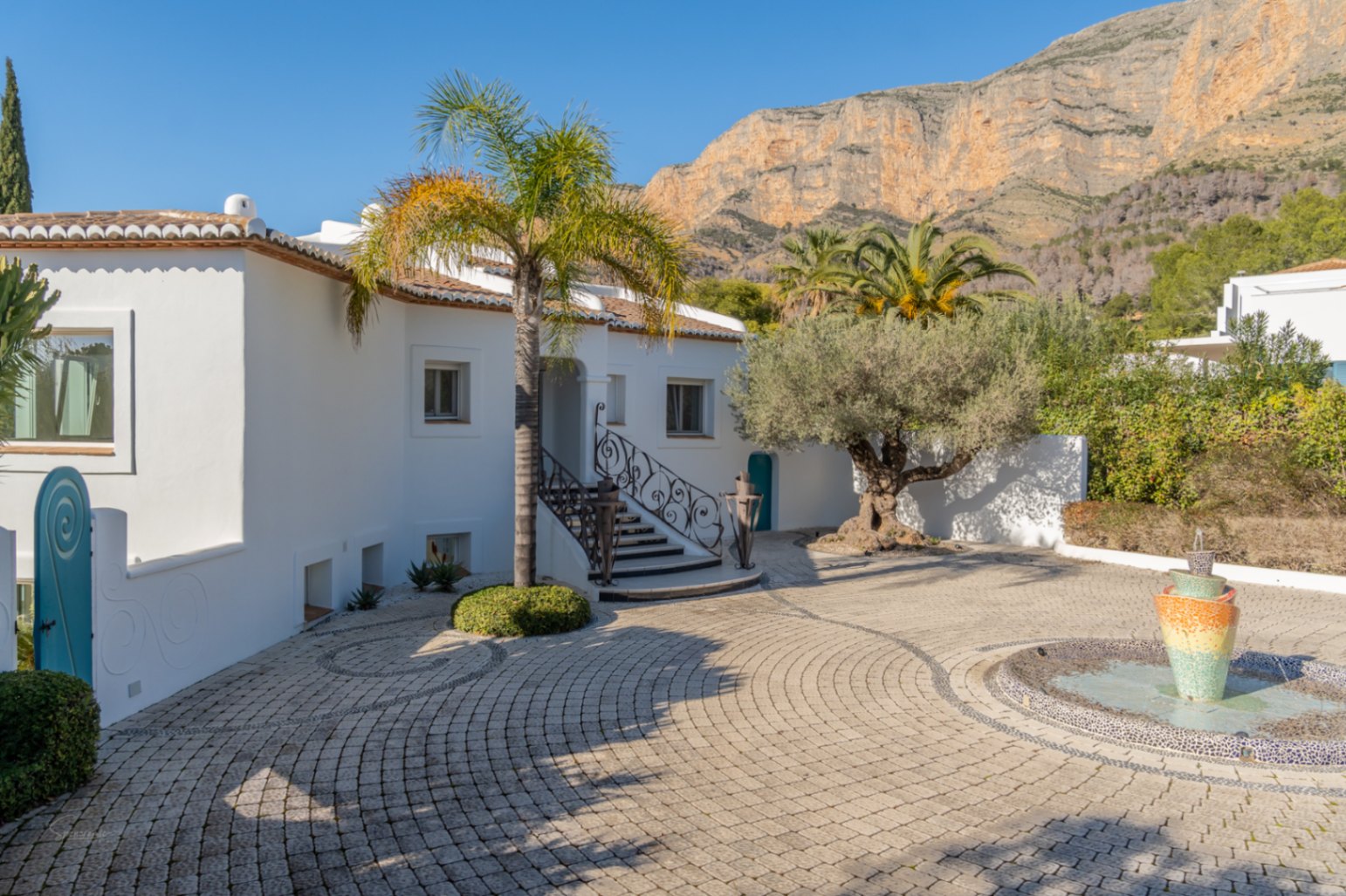 Entrada principal con patio pavimentado, fuente decorativa y palmeras, destacando la fachada blanca de estilo ibicenco bajo la majestuosa montaña del Montgó.