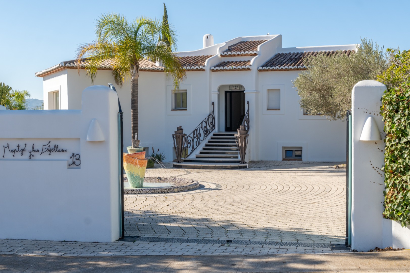 Entrada principal de la villa con fachada blanca, escalera con barandilla de hierro forjado y una fuente de mosaico sobre un patio pavimentado circular.