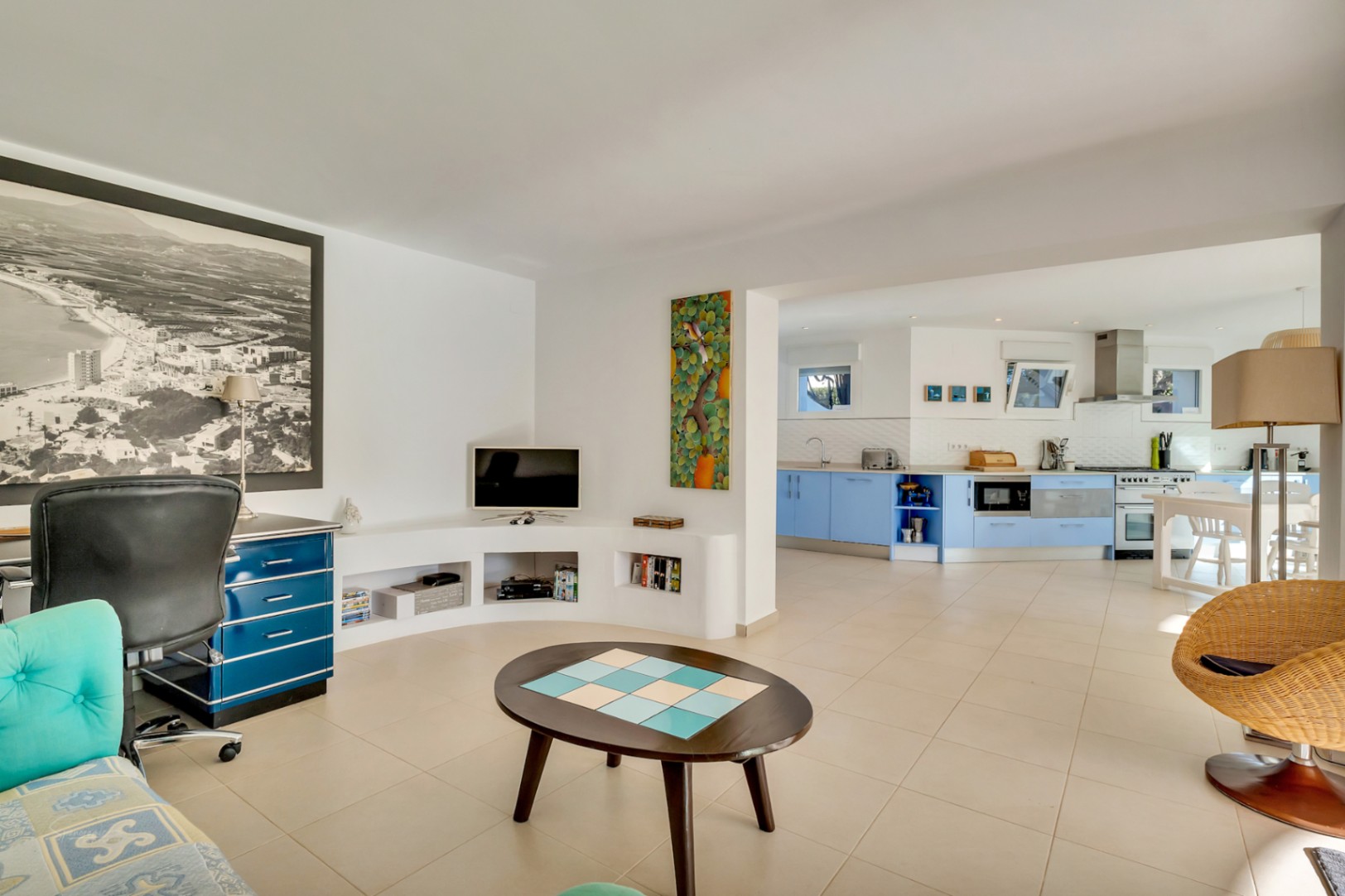 Espacio diáfano que integra una zona de oficina con escritorio azul y una cocina moderna de estilo mediterráneo con armarios en azul celeste y suelos cerámicos.