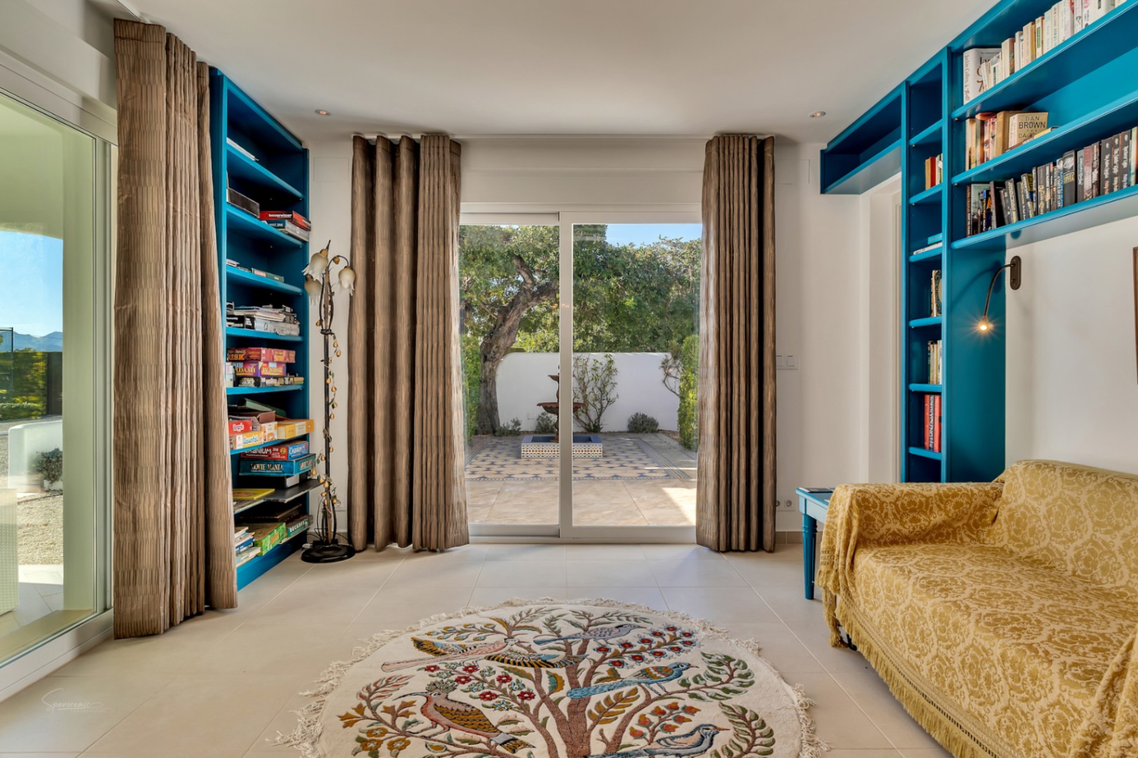 Estudio luminoso con estanterías integradas en azul vibrante, sofá tapizado en amarillo y acceso directo a un patio privado con fuente y suelos decorativos.