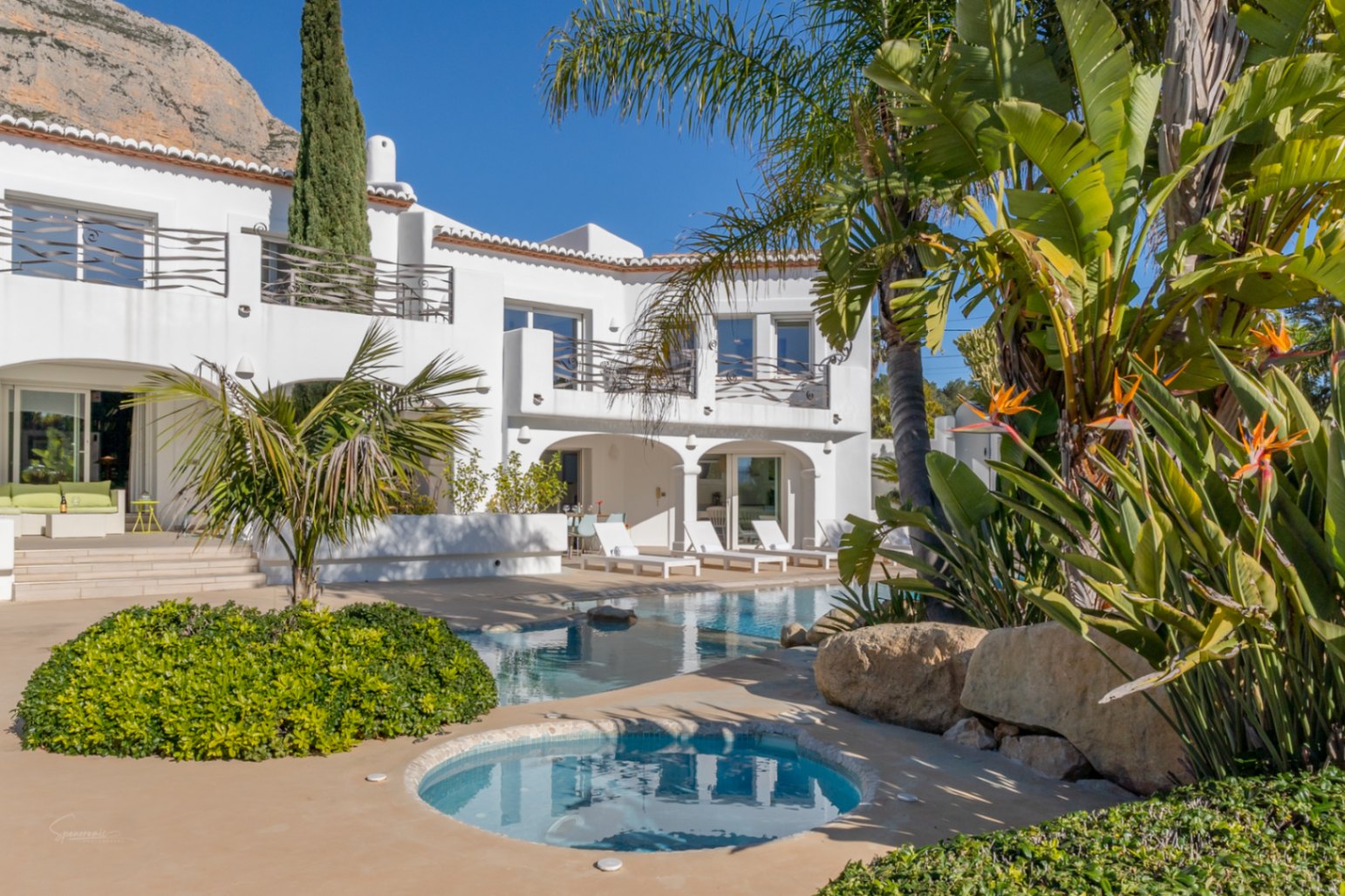 Exterior de la villa estilo Ibiza con piscina de diseño, jacuzzi integrado y naya arqueada bajo el telón de fondo de la montaña Montgó y vegetación tropical.