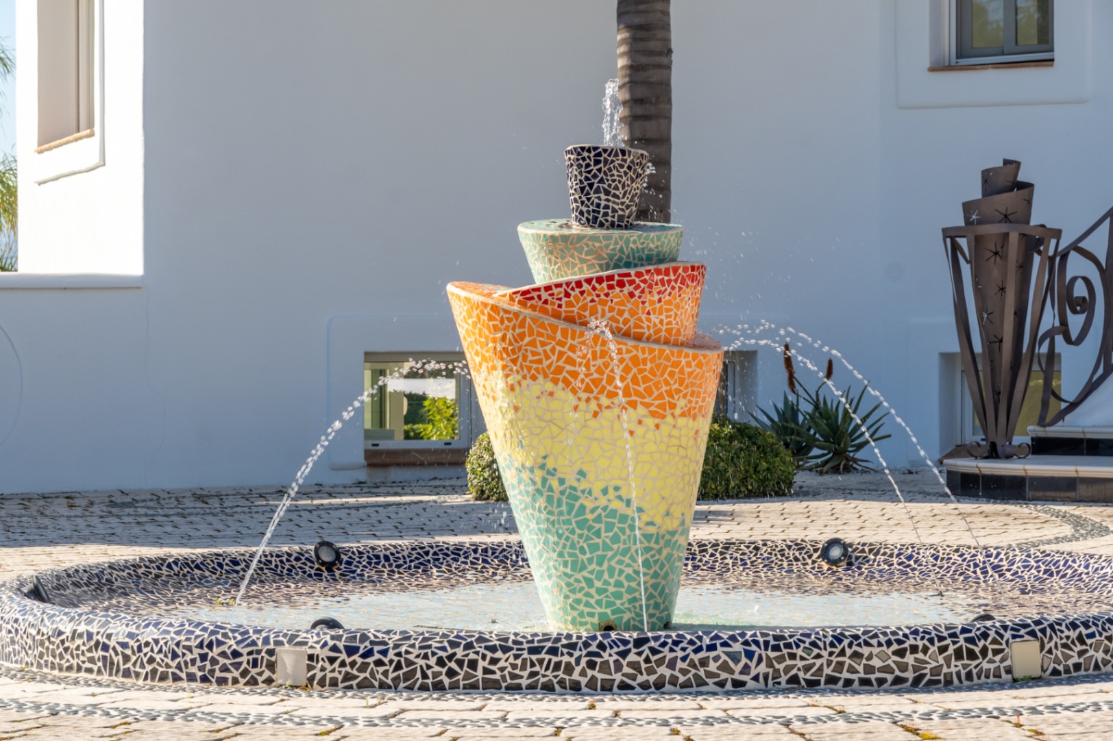 Fuente central de mosaico multicolor con diseño en espiral, situada en el patio de entrada pavimentado frente a la fachada blanca de la villa.