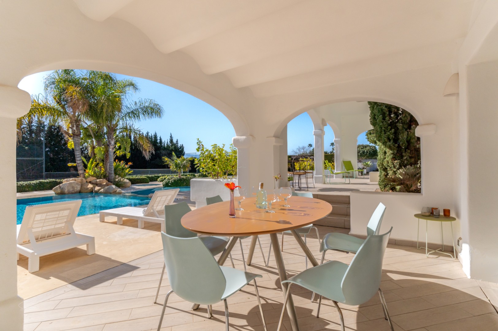 Naya cubierta con arcos blancos y comedor exterior, con vistas directas a la piscina de 70m², zona de hamacas y la pista de tenis privada rodeada de palmeras.