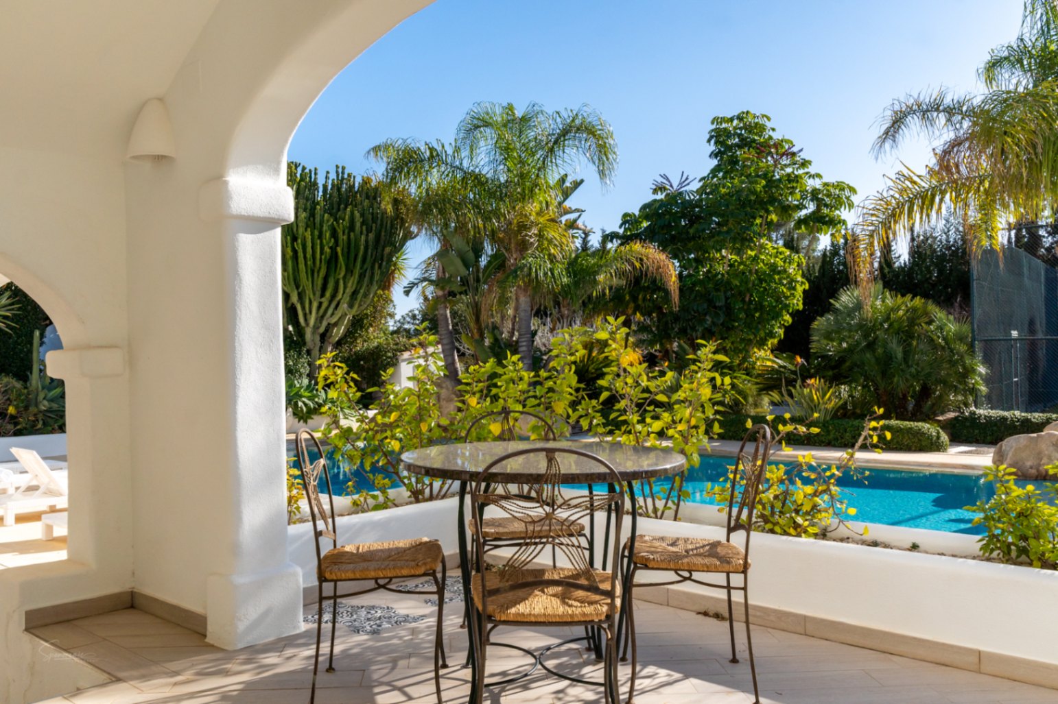 Naya de estilo ibicenco con arcos blancos, amueblada con mesa de cristal y sillas de forja, con vistas directas a la piscina privada y al jardín tropical.