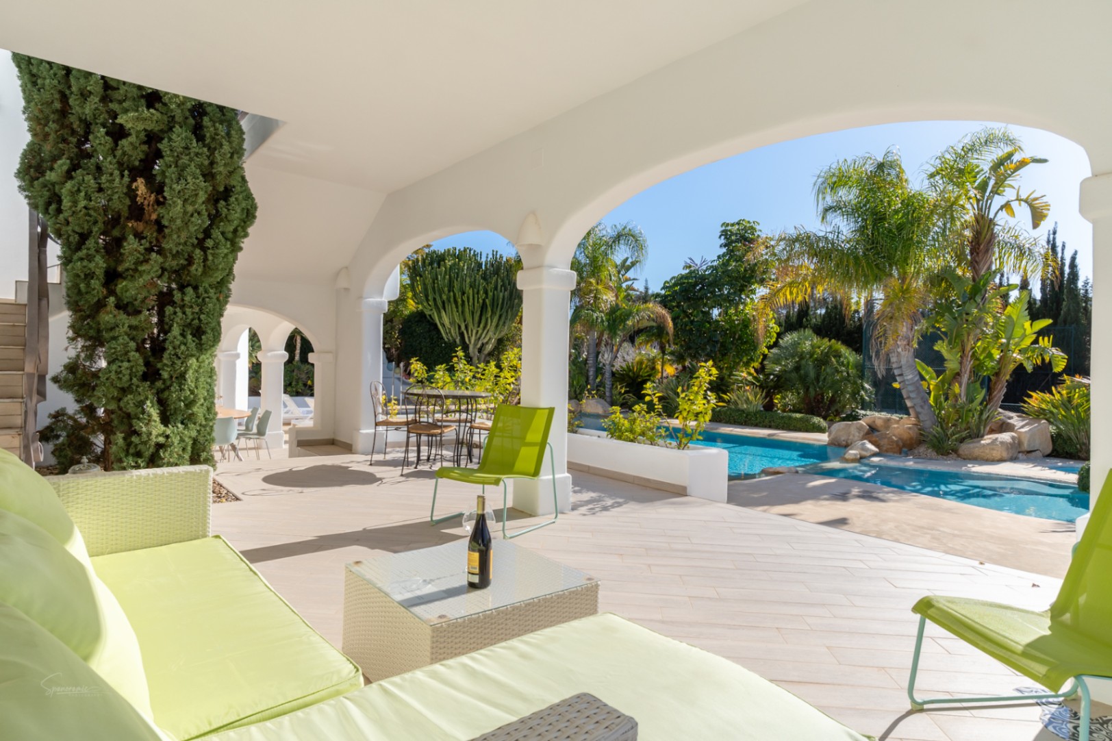 Naya sombreada con arcos blancos y mobiliario en tonos lima, con vistas directas a la piscina y al jardín mediterráneo con palmeras.