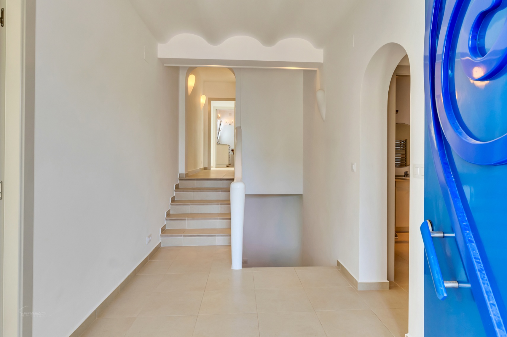 Pasillo de entrada con una llamativa puerta azul de diseño circular, suelos de gres y una escalera que conecta los niveles bajo un techo con detalles ondulados.